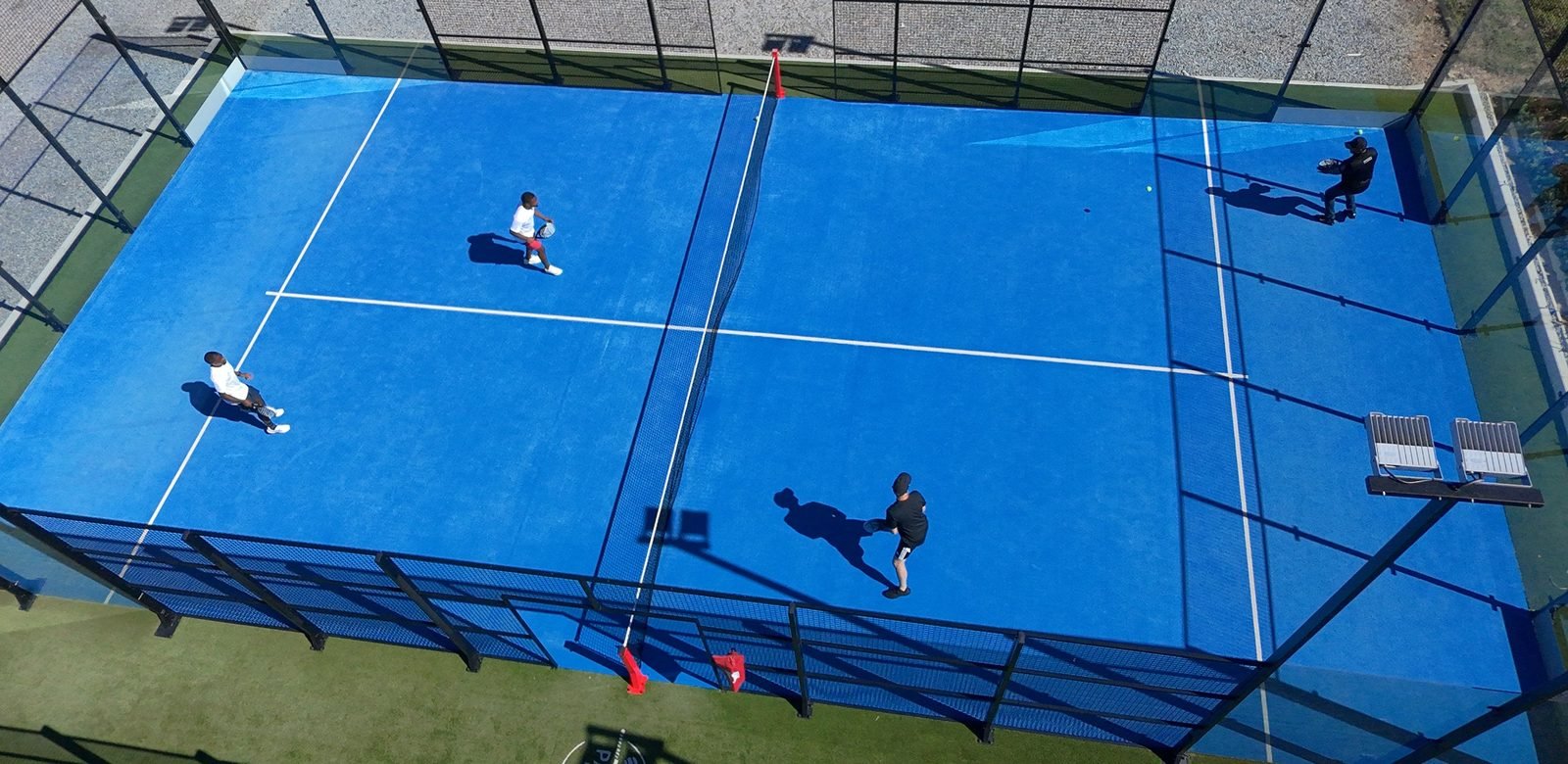 A padel court seen from directly above: four players mid-game inside the glass-and-mesh box, the net across the middle, floodlights on the side wall and long shadows across the surface.